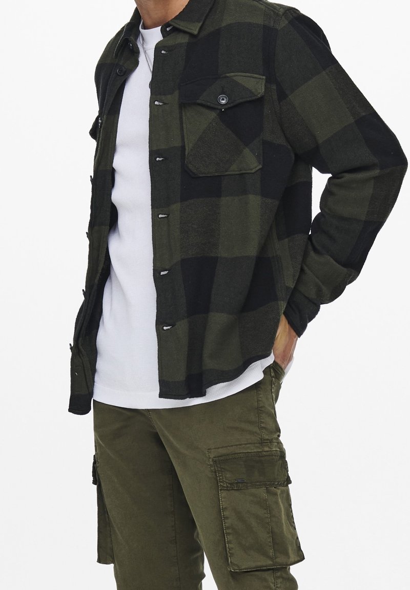 Zielono-czarna kraciasta koszula-kurtka z dwoma przednimi kieszeniami, zapinana na guziki, wykonana z teksturowanej tkaniny. W zestawie z oliwkowymi spodniami cargo.