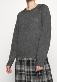 Pull en tricot gris avec un col rond, des poignets et un ourlet côtelés, porté sur une jupe à carreaux noir et blanc. Texture douce, coupe décontractée.