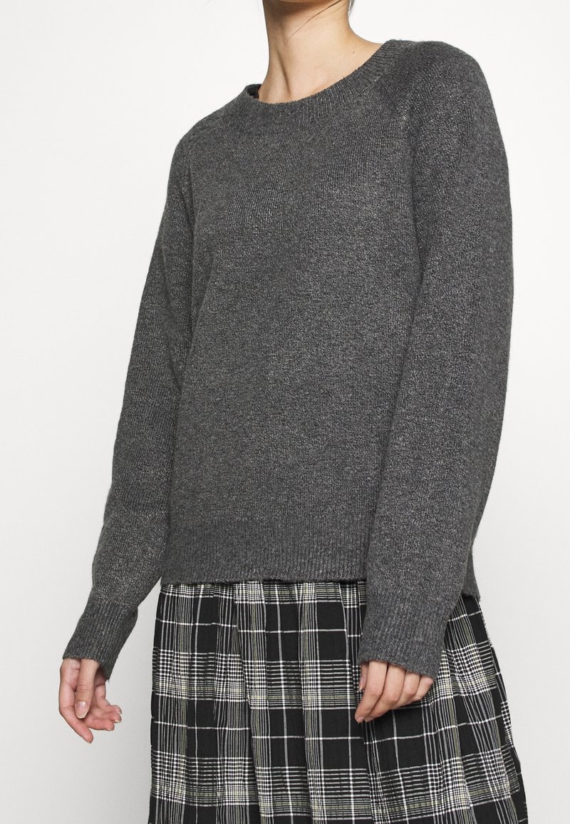 Pull en tricot gris avec un col rond, des poignets et un ourlet côtelés, porté sur une jupe à carreaux noir et blanc. Texture douce, coupe décontractée.