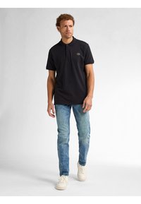 Camisa polo negra de manga corta, con un pequeño logo verde, combinada con unos vaqueros descoloridos de un azul claro y zapatillas blancas. Diseño sin costuras.
