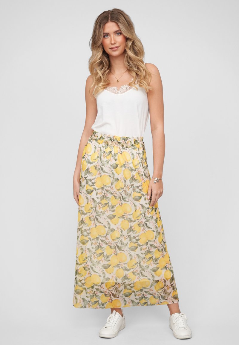 Femme blonde portant un haut blanc sans manches en dentelle, une longue jupe fleurie avec des citrons jaunes et des baskets blanches, debout devant un fond gris.