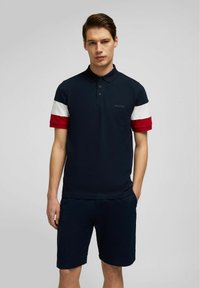 Marineblaues Poloshirt mit rot-weißen, gestreiften Kurzärmeln, zwei Knöpfen und gesticktem Logo auf der Brust. Kombiniert mit marineblauen Shorts.