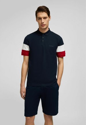 Marineblauwe poloshirt met rood en witte gestreepte korte mouwen, twee knopen met een knoopsluiting en geborduurd logo op de borst. Gecombineerd met marineblauwe korte broek.