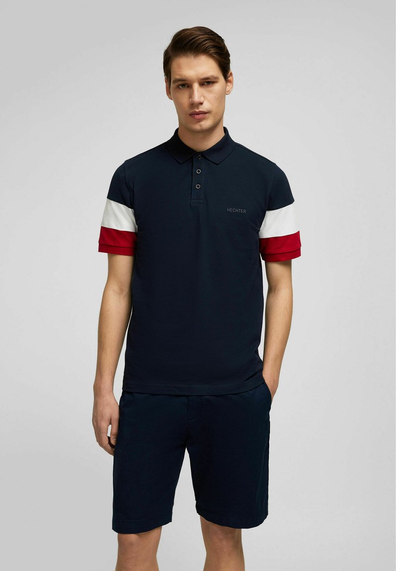Marineblaues Poloshirt mit rot-weißen, gestreiften Kurzärmeln, zwei Knöpfen und gesticktem Logo auf der Brust. Kombiniert mit marineblauen Shorts.