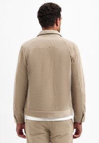 Beige corduroyjas met een gespreide kraag, zichtbare textuur en knoopmanchetten, gezien vanuit de achterkant.