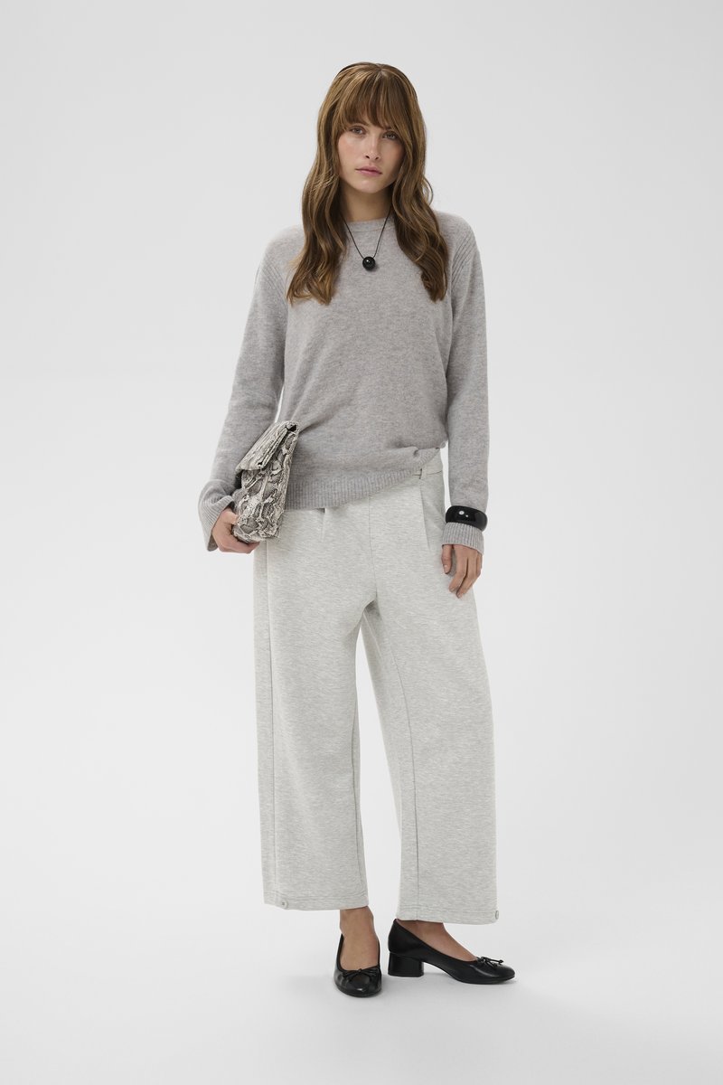 Femme debout, aux longs cheveux bruns, portant un pull gris, un pantalon large gris clair, des chaussures noires à talons bas, et tenant une pochette texturée.