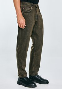Jeans marrones oscuros con una tela texturizada, corte recto, diseño de cinco bolsillos y una pequeña etiqueta de marca roja cerca del bolsillo trasero. Llevados con botas negras.