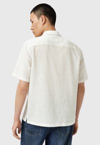 Chemise blanche à manches courtes avec un tissu texturé, coupe décontractée et col replié. Le dos présente une subtile couture et des fentes sur les côtés.