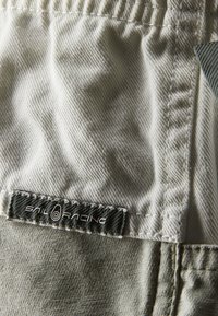 Beige tyg med texturerad yta, med en grå etikett som läser "SAIL RACING" och är fäst vid sömmen.