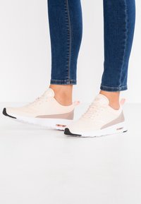 Light pink athletic sneakers with a textured mesh upper, contrasting beige accents, white sole, and black tip. Features laces and a pull tab.