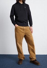 Persona in felpa nera Puma, pantaloni in corda beige a vita alta e scarpe in pelle marrone, in piedi su un tappeto blu contro un muro bianco.