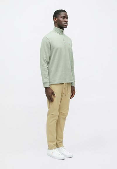 Πράσινη, μακρυμάνικη φούτερ με φερμουάρ και υφή συνδυασμένων υλικών, σε συνδυασμό με μπεζ cargo παντελόνι και λευκά αθλητικά παπούτσια. Απλός σχεδιασμός με ελάχιστες λεπτομέρειες.
