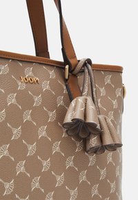 Sac cabas marron texturé avec un motif blanc répétitif, ornée d'un logo doré et de pompons décoratifs assortis en matériau similaire.