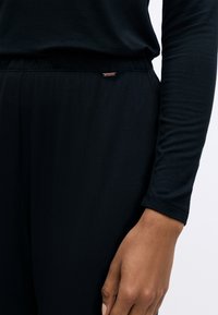 Črna dolgosleeve majica z gladko teksturo, ki ima rahlo sijaj in majhen kovinski logotip na pasu. Povečan pogled na rokav in pas.