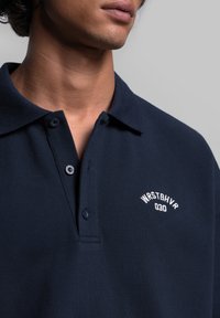 Granatowa koszulka polo wykonana z teksturowanej tkaniny z dwoma guzikami. Posiada białe haftowane logo na lewej piersi.