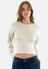 Maglione bianco a costine con scollatura rotonda e maniche lunghe, caratterizzato da una texture lucida. Indossato con jeans aderenti blu scuro.