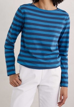 Personne portant une chemise à manches longues rayée bleu et marine associée à un pantalon blanc, debout avec une main dans la poche.