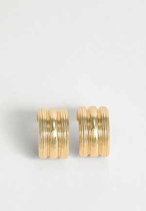 Paire de grandes boucles d'oreilles créoles dorées avec un design strié superposé sur un fond neutre.