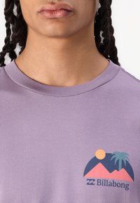 T-shirt en coton violet avec un imprimé graphique de montagnes, un soleil et un palmier en rouge, bleu et jaune près de la zone gauche de la poitrine.