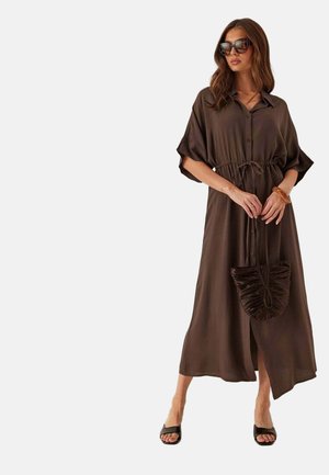 Donna che indossa un vestito midi marrone con bottoni e cintura in vita, sandali neri, occhiali da sole e tiene in mano una borsa marrone arricciata abbinata.