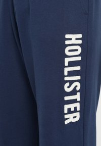 Tengerész kék szövet, fehér hímzett betűkkel, amelyek függőlegesen írják a "HOLLISTER" szót egy varrás mentén.