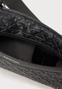 Sort lædertaske med en struktureret, præget geometrisk mønster, lynlås lukning og en glat indvendig foring. Mærkenavn synligt indeni.