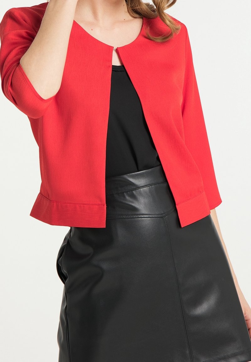 Femme portant un cardigan court rouge par-dessus un haut noir rentré dans une jupe en cuir noire taille haute, visible des épaules jusqu'au milieu des cuisses.