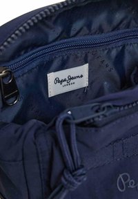 Interno di una borsa blu navy con tessuto texturizzato, dotato di una tasca con zip e un'etichetta che recita "Pepe Jeans London." Cuciture dettagliate visibili.