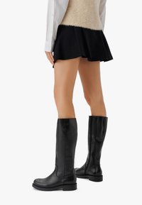 Osoba v černých kožených holínách po kolena, krátké černé sukni, bílém tričku a béžovém bezrukávovém svetru, stojící na bílém pozadí.