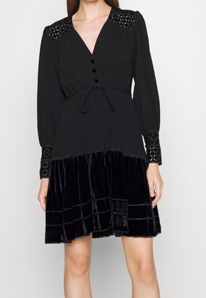 Czarna sukienka z długim rękawem, dekoltem w serek, guzikami, paskiem w talii, zdobionymi ramionami i mankietami oraz aksamitnym materiałem spódnicy sięgającej nad kolana.