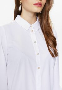 Camicia bianca a maniche lunghe con colletto classico e dettagli di bottoni perlacei, caratterizzata da una texture liscia e una tasca singola sul lato sinistro.