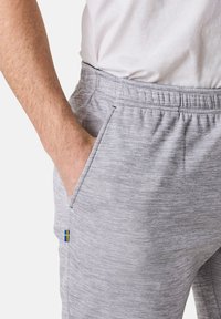 Grau sportliche Hosen mit einem elastischen Bund, seitlichen Taschen und einem kleinen blau-gelben Emblem der schwedischen Flagge. Strukturierte Stoffdetails sichtbar.
