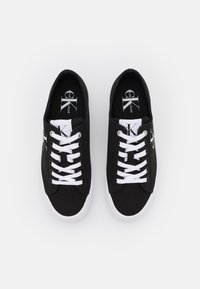 Svarta kanvas sneakers med vita snören och sulor. Har en logotyp på sidan och en platt tådesign. Slät textur och vadderad insida.