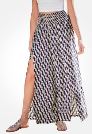 Vrouw draagt een lange rok met patroon en een hoge split, samen met platte sandalen met decoratieve bandjes, gezien van taille tot voeten.