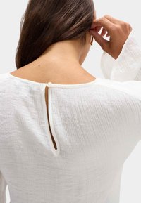 Blusa bianca con texture, scollatura rotonda e chiusura a bottoni posteriori, realizzata in tessuto leggero e crease, con maniche lunghe.