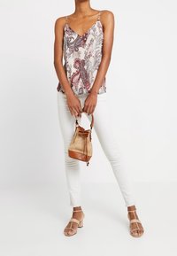 Kvinna som bär en ärmlös blus med paisleytryck, vita skinny jeans, beiga klackar med remmar och håller en tan vävd handväska med läderdetaljer.