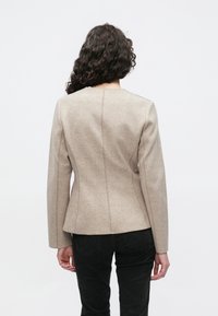 Beige op maat gemaakte blazer van gestructureerde stof met een getailleerd silhouet en lange mouwen. Achteraanzicht toont strakke naden en minimale details.