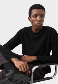 Pull en maille noire à col rond, porté par une personne assise sur une chaise chromée. Caractérisé par une texture lisse et des manches ajustées, associé à un pantalon habillé.