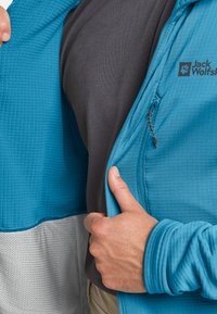 Giacca blu testurizzata con accenti grigi; strato interno nero visibile. Il tessuto presenta un motivo a griglia e include una zip con linguetta.