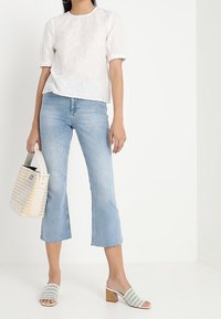 Biała bluzka z bufkami, jasnoniebieskie dzwony, białe mułowate buty na obcasie z odkrytymi palcami i perforowanym wzorem, trzymając torebkę w odcieniu kremowym o teksturowanej powierzchni.