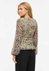 Blondide juustega naine, kes kannab beeži ja musta mustriga pika varrukaga pluusi, millel on volangvarrukad, ja musti pükse, vaadates üle oma õla.