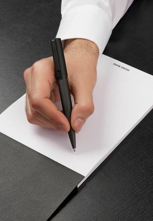 Černé texturované pero držené v ruce, píšící na prázdném bílém notýsku, umístěném na tmavém dřevěném povrchu.