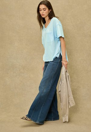 Mladá žena ve světle modrém tričku s výstřihem do V a širokých džínách, držící v ruce proužkované tričko s dlouhým rukávem, na béžovém pozadí.