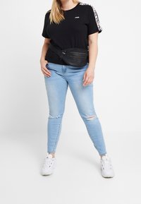 Černé tričko s logem, modré seprané skinny džíny a černá ledvinka. Bílé tenisky dotvářejí neformální outfit.