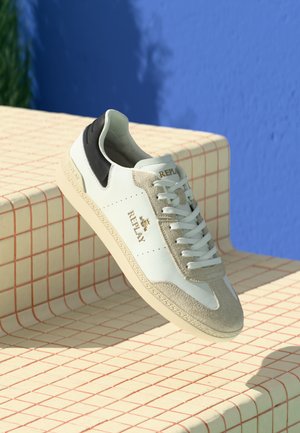 Sneaker Replay bianca e beige con tallone nero, lacci bianchi e logo dorato appoggiata su una superficie piastrellata crema contro un muro blu.