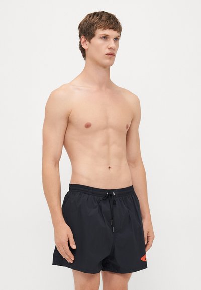 Ein junger Mann mit kurzen Haaren, der schwarze Schwimmshorts mit Kordelzug trägt, steht vor einem schlichten weißen Hintergrund und schaut zur Seite.