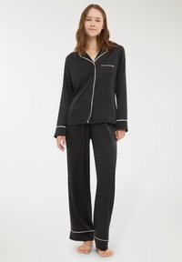 Giovane donna in piedi a piedi nudi, indossando un completo pigiama nero a maniche lunghe con dettagli in bianco sui polsini e sul colletto.