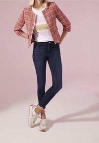 Vrouw draagt een roze geruite korte jas, wit grafisch t-shirt, donkere skinny jeans, beige sokken en witte sneakers op een roze achtergrond.