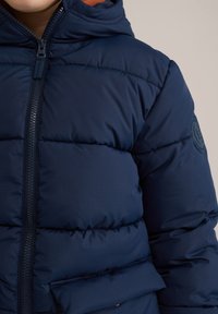 Tamsiai mėlynas pūkinis švarkas su užtrauktuku priekyje, kapišonu ir grublėta tekstūra. Yra siuvinėti paneliai ir apvalus logotipo ženkliukas ant rankovės.