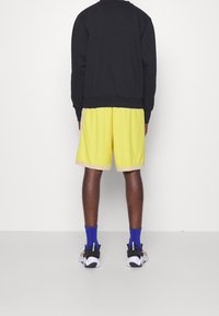 Sudadera negra, pantalones cortos amarillos con ribete beige, calcetines azules y zapatillas. Diseño simple con un corte relajado y un contraste de colores vibrante.